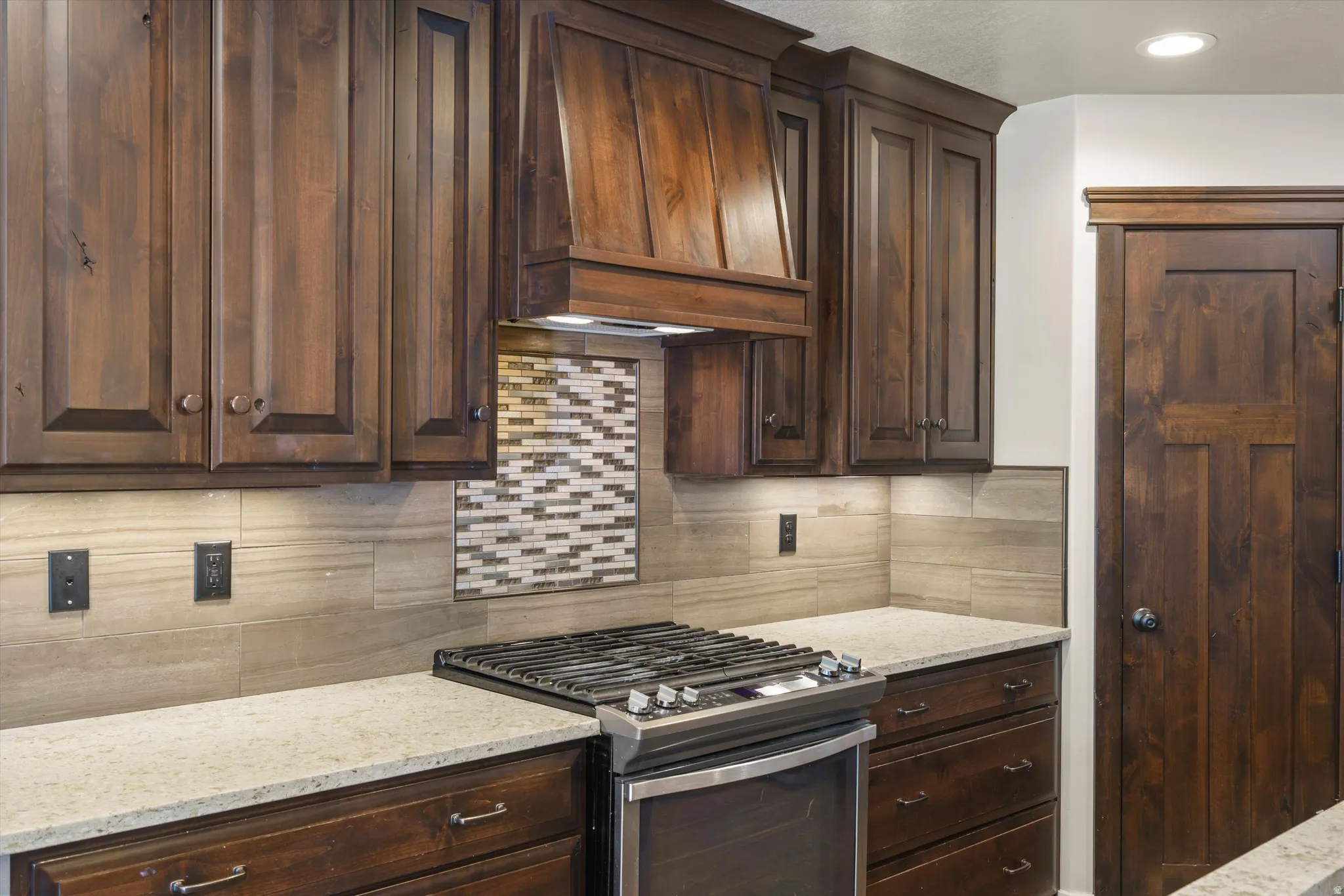 Wood cabinetry with integrated range hood, built-in cooktop, light-colored countertops, and tiled backsplash