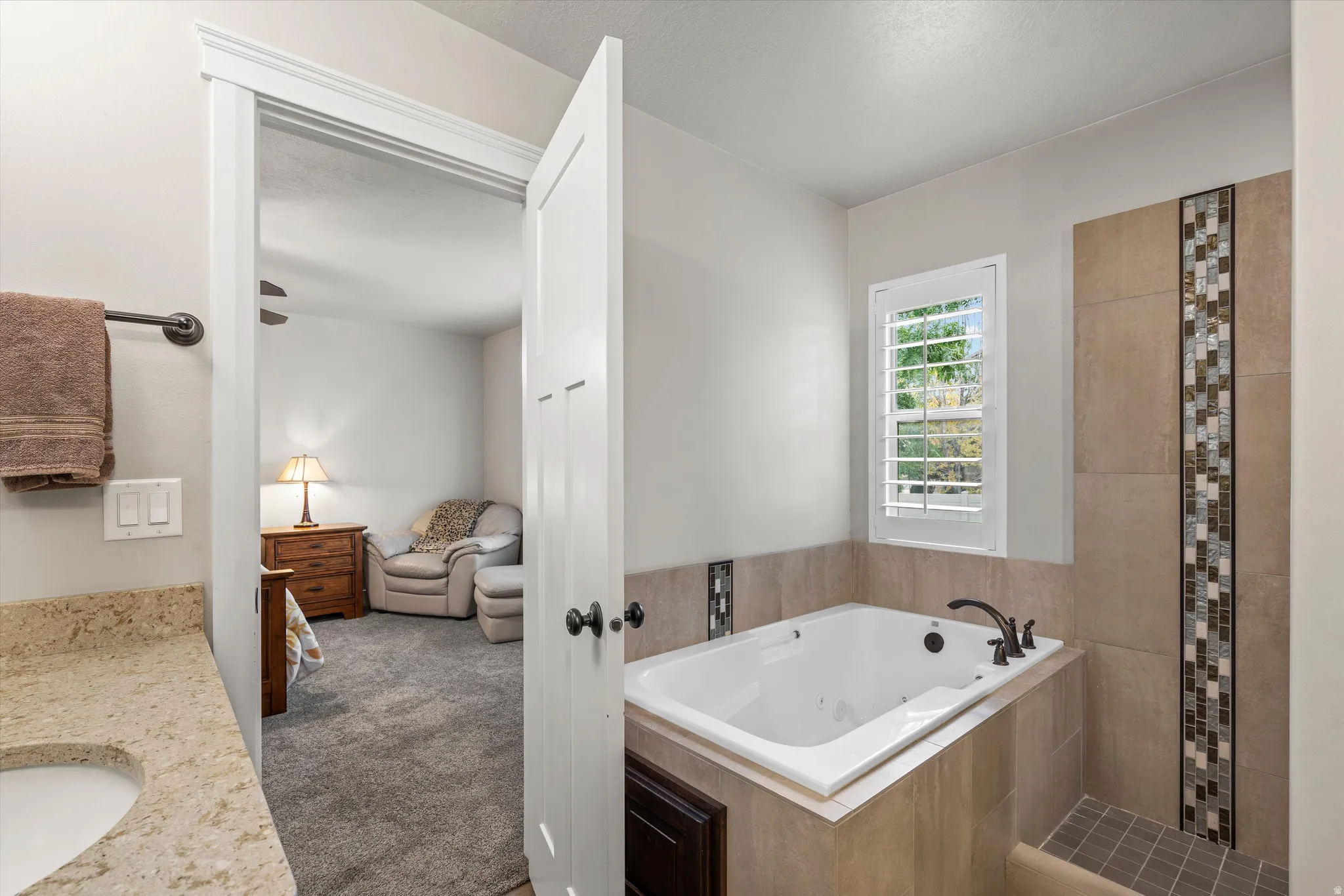 Built-in jetted tub, separate tiled shower with mosaic accent, window with plantation shutters, vanity with stone countertop and undermount sink