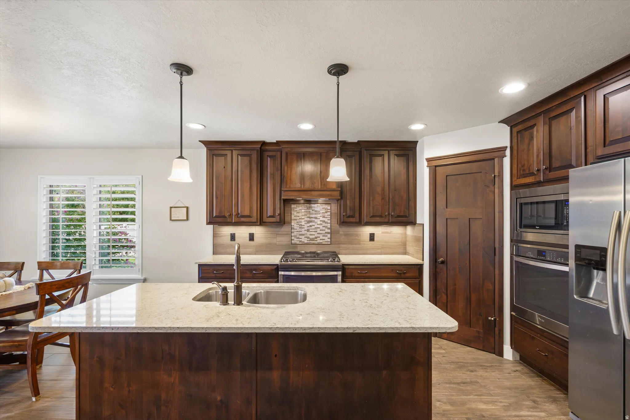 Kitchen featuring dark wood cabinetry, a center island with a double basin sink and light-toned countertop, stainless steel appliances, and pendant lighting