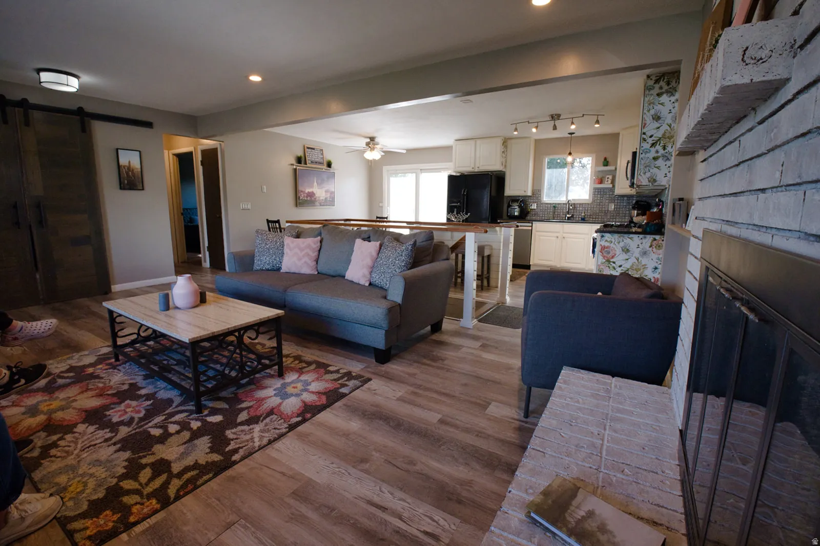 Open-concept living area featuring wood-finish flooring, a painted brick fireplace, and a kitchen with white cabinetry and dark appliances