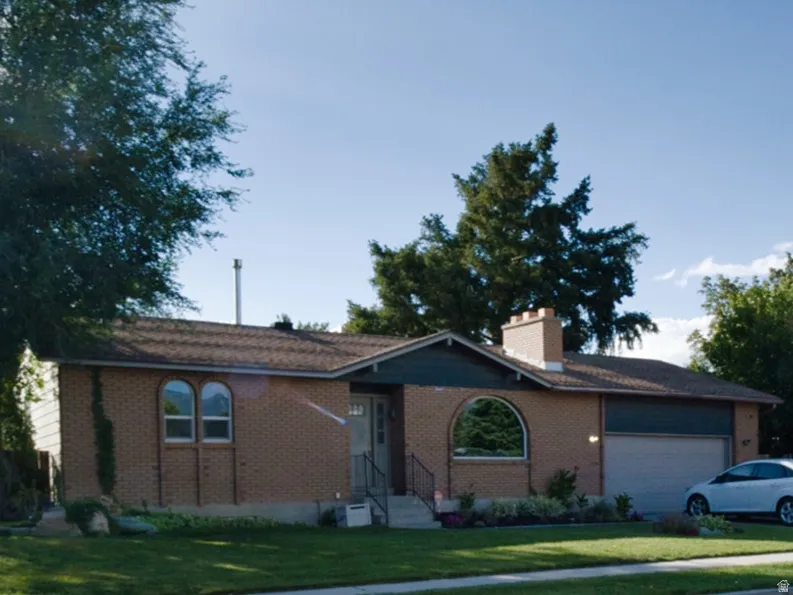 Brick exterior with a covered entry, arched window, attached garage, and mature landscaping