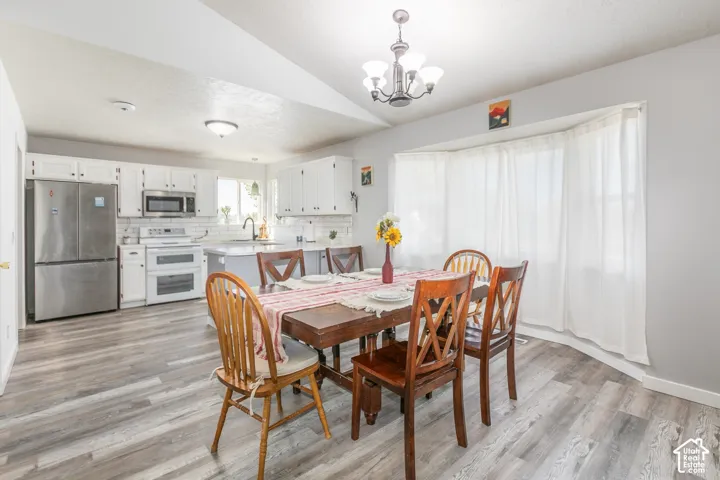 Vaulted ceiling with chandelier, wood-finish flooring, bay window, white kitchen cabinetry, and stainless steel appliances