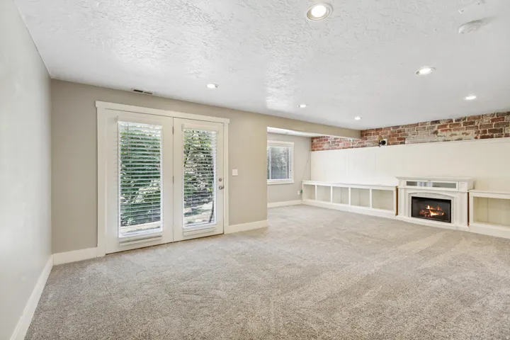Recessed lighting, exposed brick accent wall, built-in shelving, white fireplace surround, and French doors with blinds