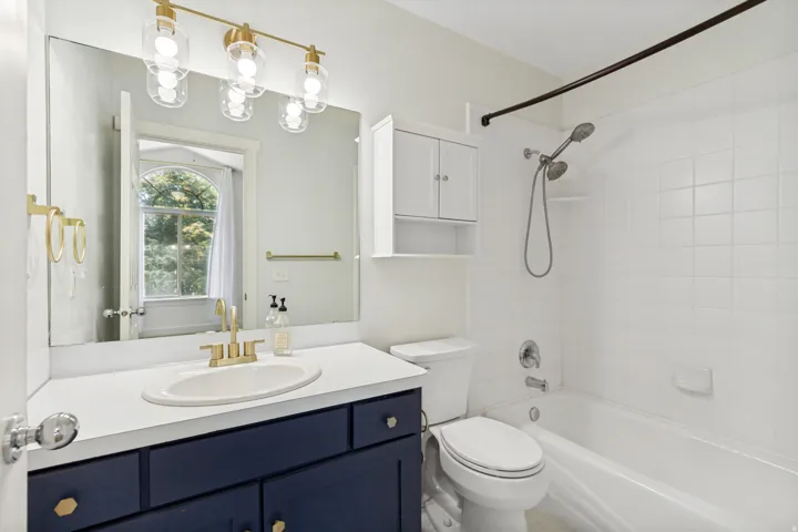 Vanity with a white countertop and navy cabinetry, gold-finish fixtures, large frameless mirror, and a built-in shower/tub combination with white tile surround