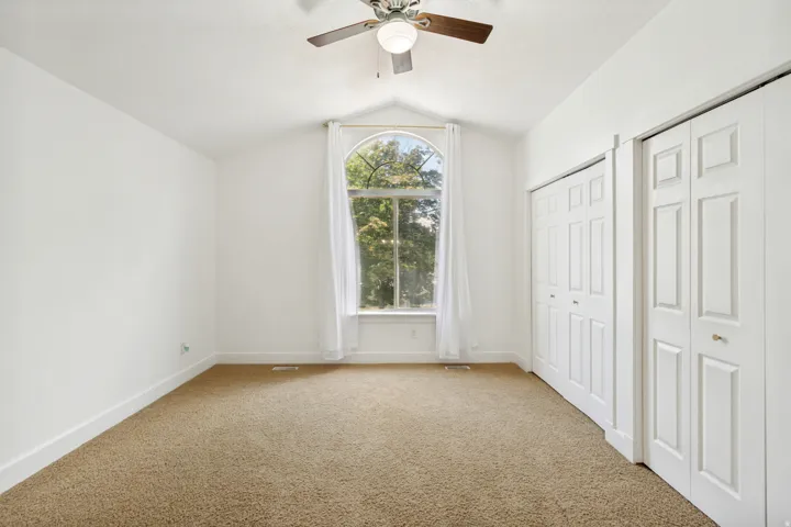 Arched window with white trim, vaulted ceiling, ceiling fan with light fixture, carpeted flooring, and bi-fold closet doors