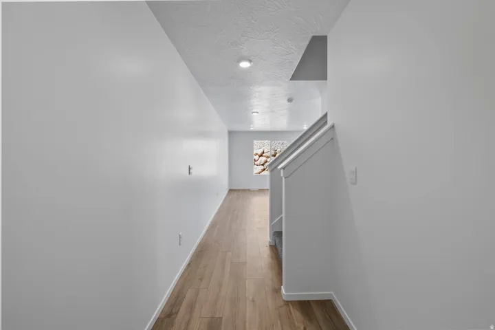 Wood-finish flooring extending through the hallway, staircase with white railing, recessed ceiling lighting, and a large window providing natural light