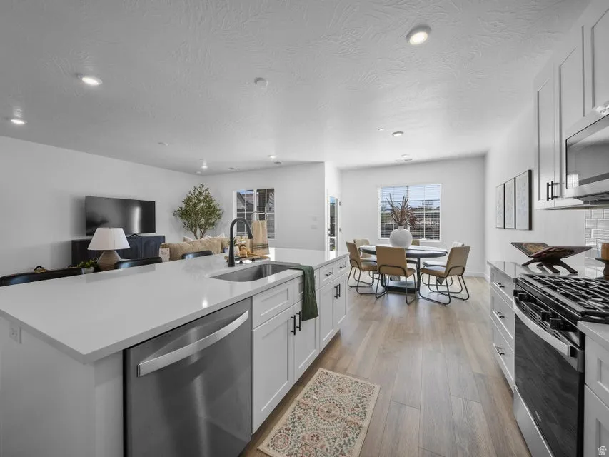 Open-concept living space featuring wood-finish flooring, recessed lighting, white cabinetry with dark hardware, a kitchen island with an undermount sink, and stainless steel appliances