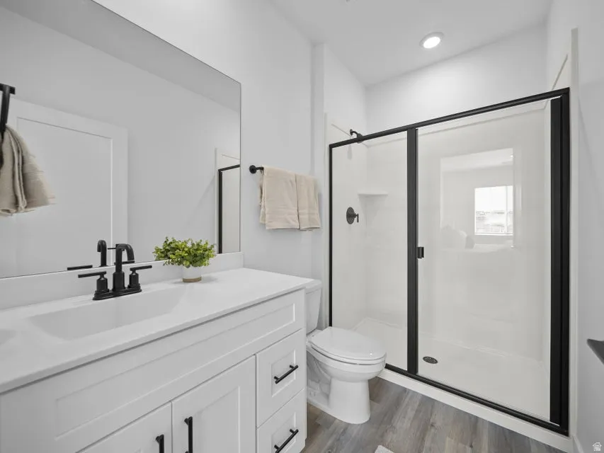 White vanity with black hardware, white countertop, rectangular undermount sink with a matte black faucet, wood-finish flooring, and a glass-enclosed shower with black framing