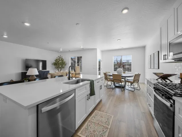 Open-concept living space featuring wood-finish flooring, recessed lighting, white cabinetry with dark hardware, a kitchen island with an undermount sink, and stainless steel appliances