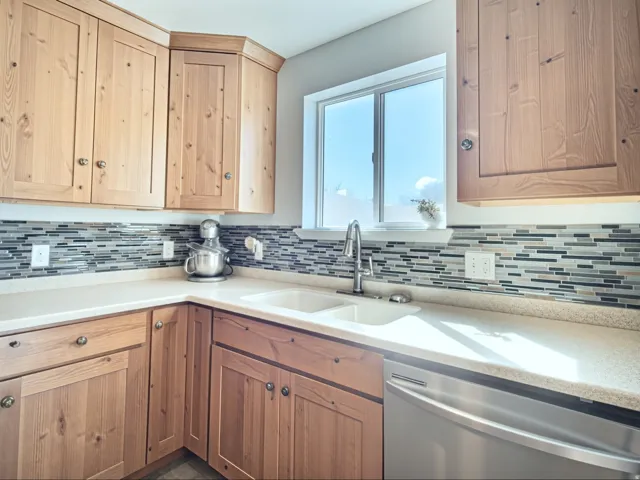 Light wood-finish cabinetry, mosaic tile backsplash, double basin sink with a high-arc faucet, durable countertop surfaces, and stainless steel dishwasher