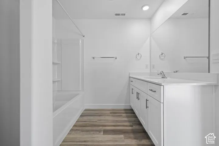 Spacious bathroom featuring a white vanity with ample storage, a large wall-mounted mirror, a tub and shower combination with white surround, and wood-finish flooring