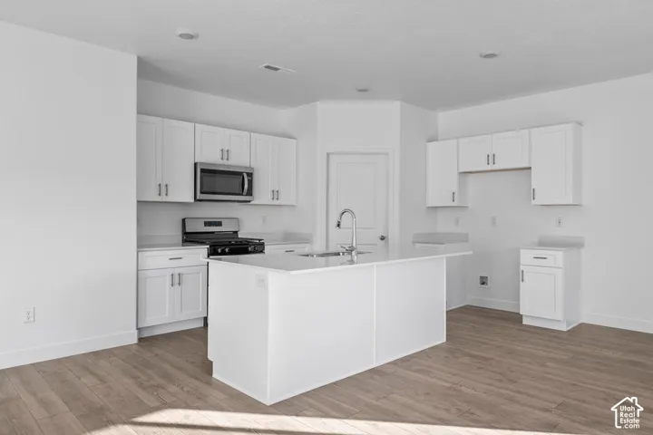 White kitchen cabinetry with stainless steel appliances, central island with a sink, light-toned wood-finish flooring, and recessed lighting