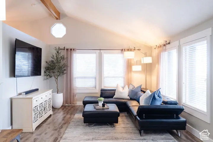 Vaulted ceiling with exposed wood beam, octagonal accent window, expansive windows with white blinds, wood-finish flooring, and a built-in media console