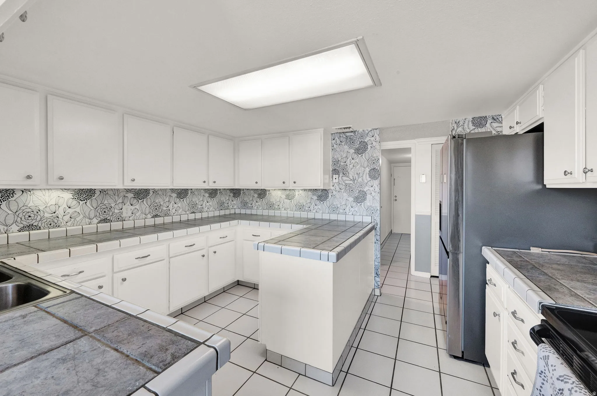 White cabinetry with brushed nickel hardware, tile countertops, stainless steel refrigerator, tile flooring, and integrated ceiling lighting