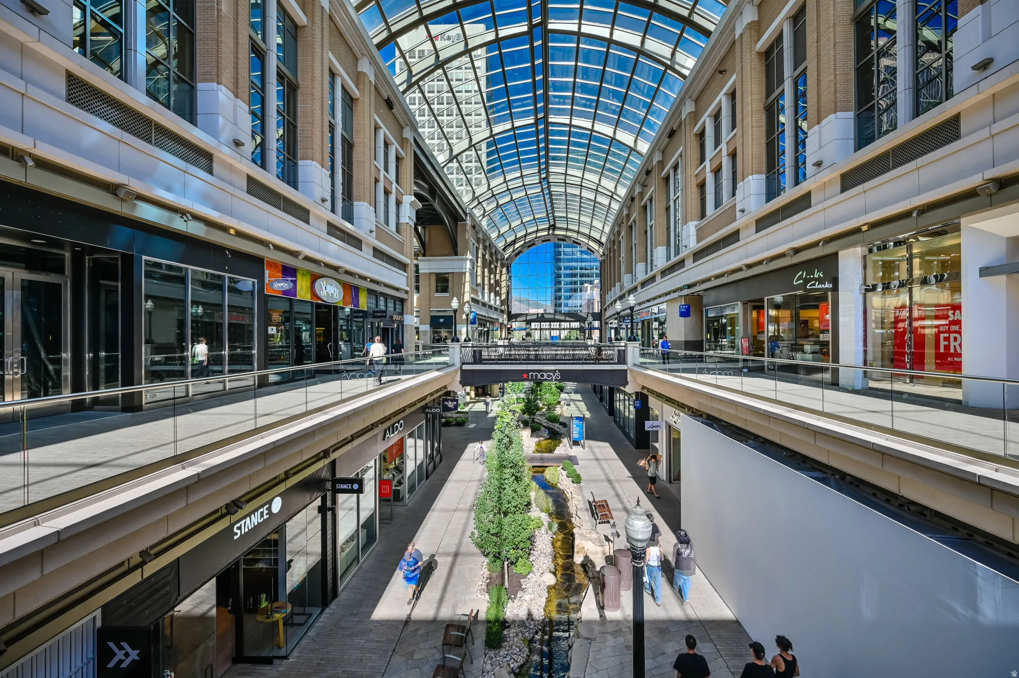 Expansive arched glass ceiling spanning multiple levels of retail space, featuring a central water feature with mature trees, and a pedestrian bridge