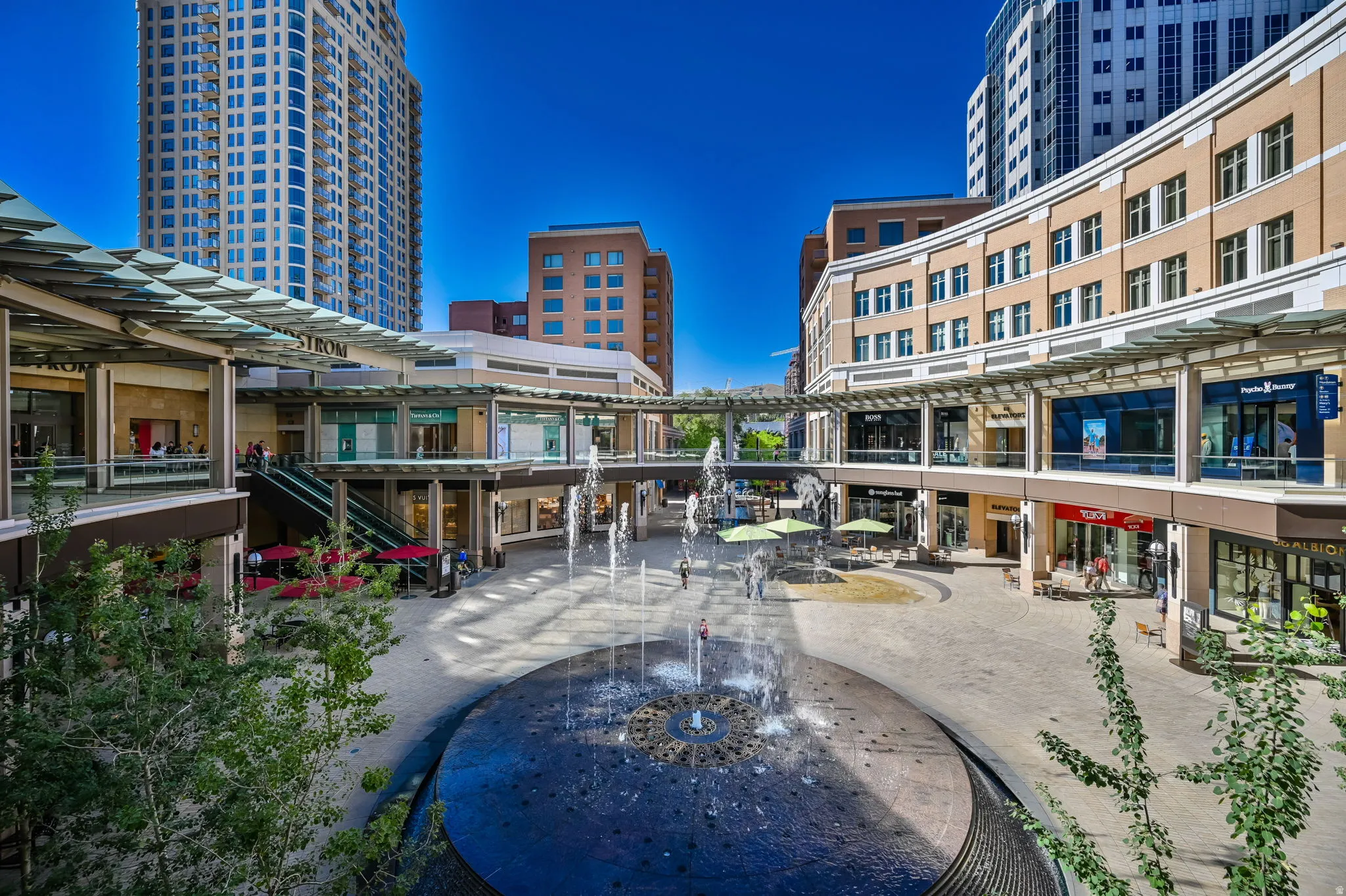 Central plaza featuring a large water fountain, tiered commercial buildings with multiple storefronts, and a contemporary high-rise structure
