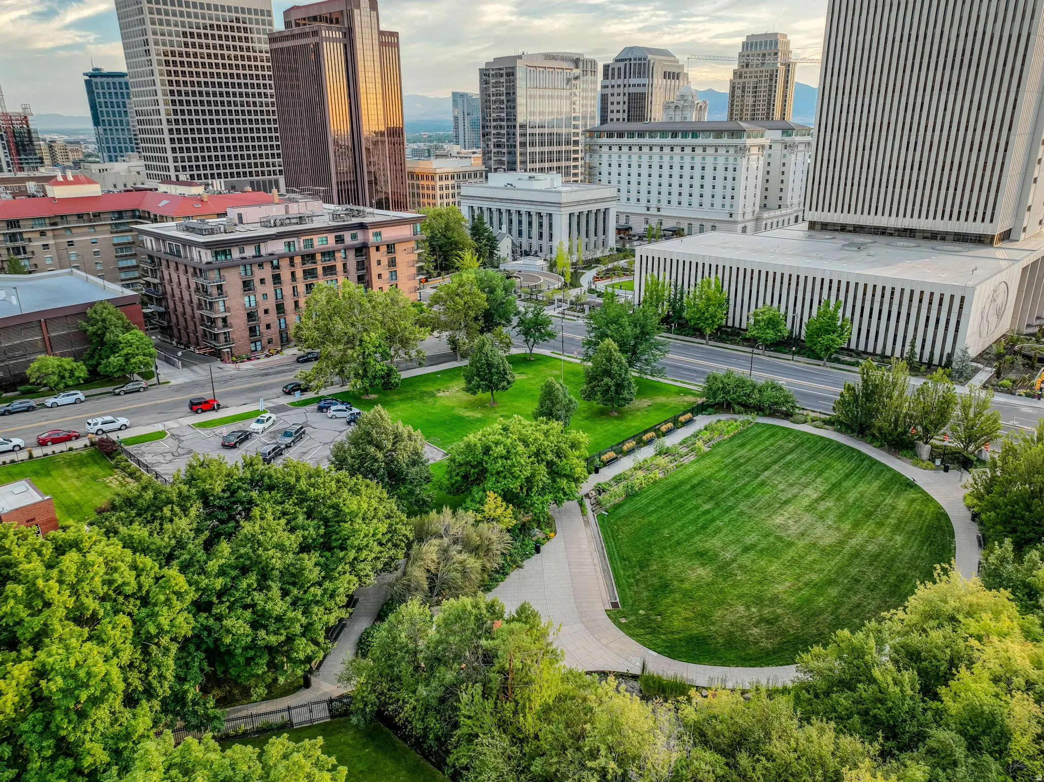 Expansive urban green space featuring a large grassy area, surrounding trees, and paved walkways, complemented by a prominent city skyline and various buildings