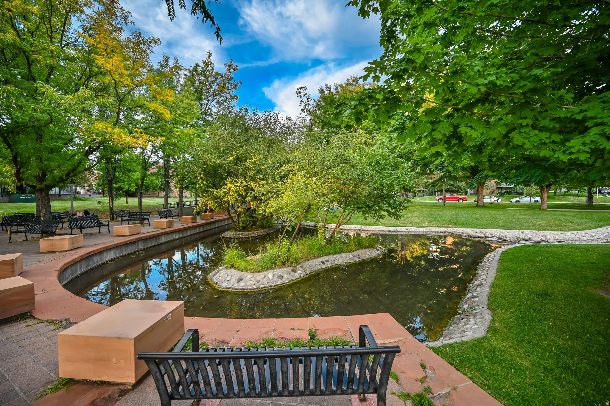 Landscaped park area featuring a pond with a central island, curved paved pathways, multiple benches, and mature trees