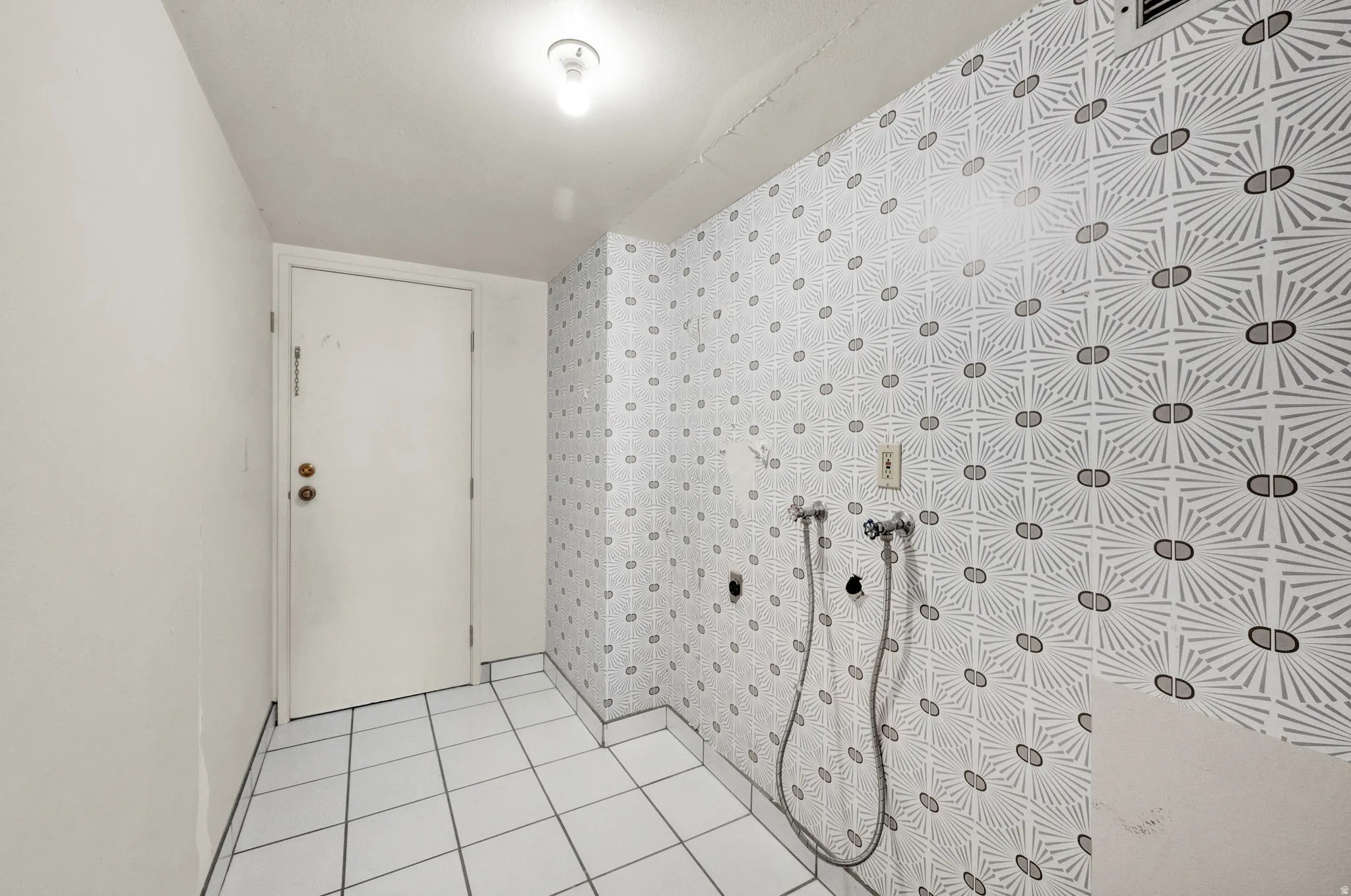 White square floor tiles, patterned wall covering, white door with brass-finish hardware, exposed utility connections
