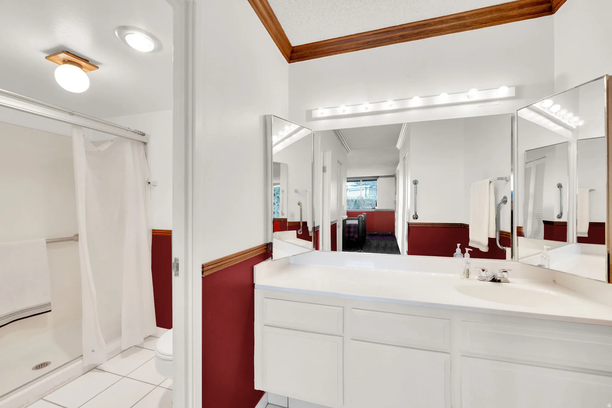 Dual vanity with white cabinetry and integrated sinks, wide wall-mounted mirror with integrated lighting, and a walk-in shower with grab bars