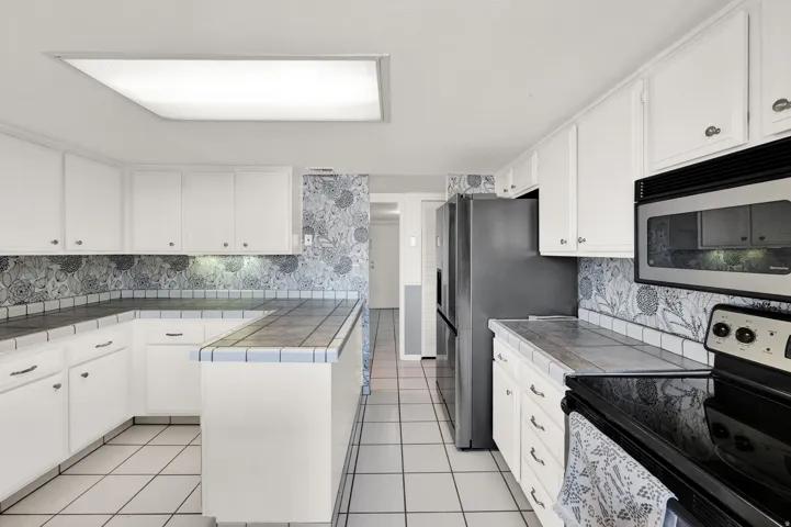 White cabinetry with dark hardware, tile flooring, built-in microwave, dark countertop, and stainless steel refrigerator