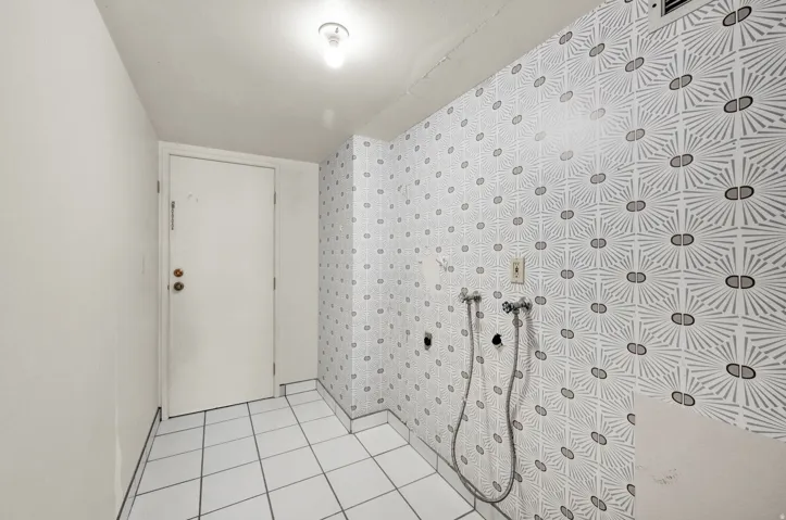 White square floor tiles, patterned wall covering, white door with brass-finish hardware, exposed utility connections