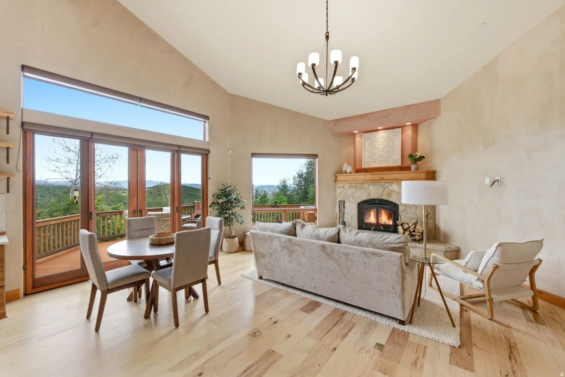 Vaulted ceiling with an exposed beam, light wood-finish flooring, stone-clad fireplace with wood mantel, large glass-paneled doors, and a deck with wood railings
