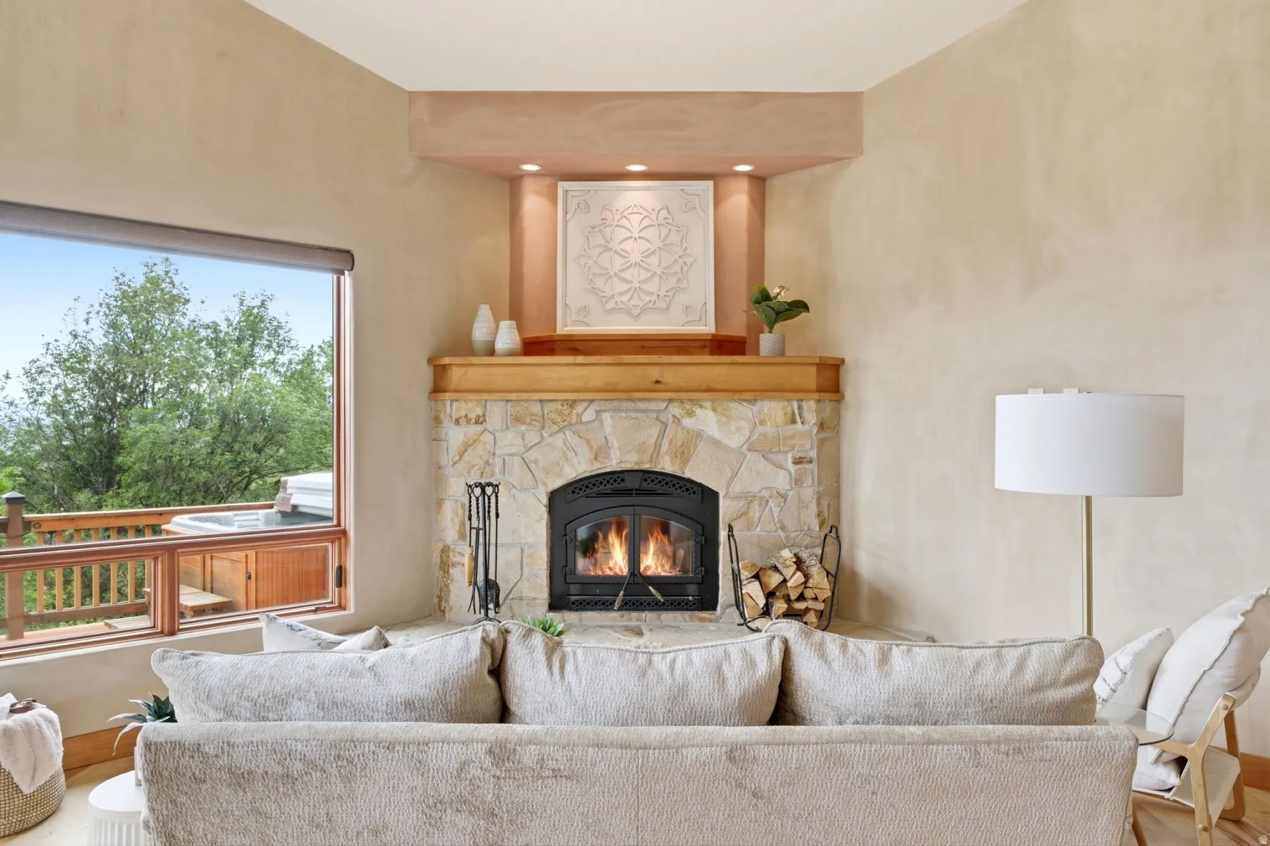 Stone-clad fireplace with a wood mantle, built-in recessed lighting, and a large window overlooking an outdoor hot tub on a wood deck