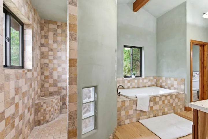 Curved stucco wall, wood-finish flooring, built-in soaking tub with tile surround, walk-in shower with tile walls and a built-in bench, and exposed wood beam ceiling