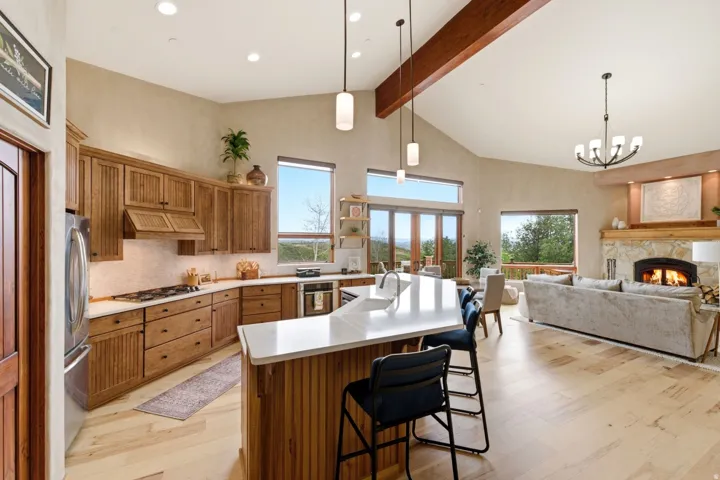 Vaulted ceilings with exposed wood beam, kitchen island with countertop seating, wood-finish cabinetry, stone fireplace with mantel, and wood-finish flooring