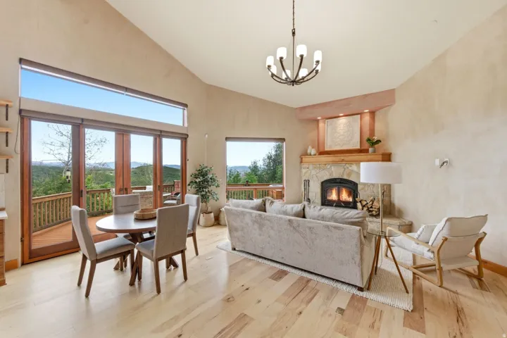 Vaulted ceiling with an exposed beam, light wood-finish flooring, stone-clad fireplace with wood mantel, large glass-paneled doors, and a deck with wood railings