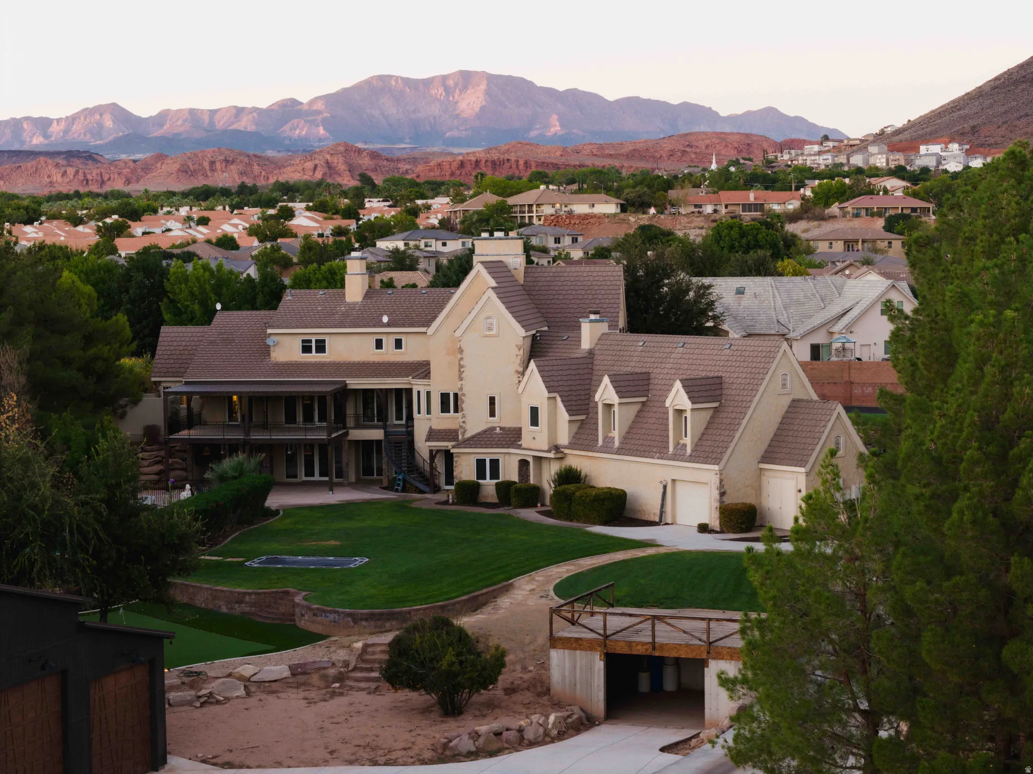 Multi-level stucco exterior with dormer windows, a covered balcony, a lower patio, a manicured lawn, and an integrated garage