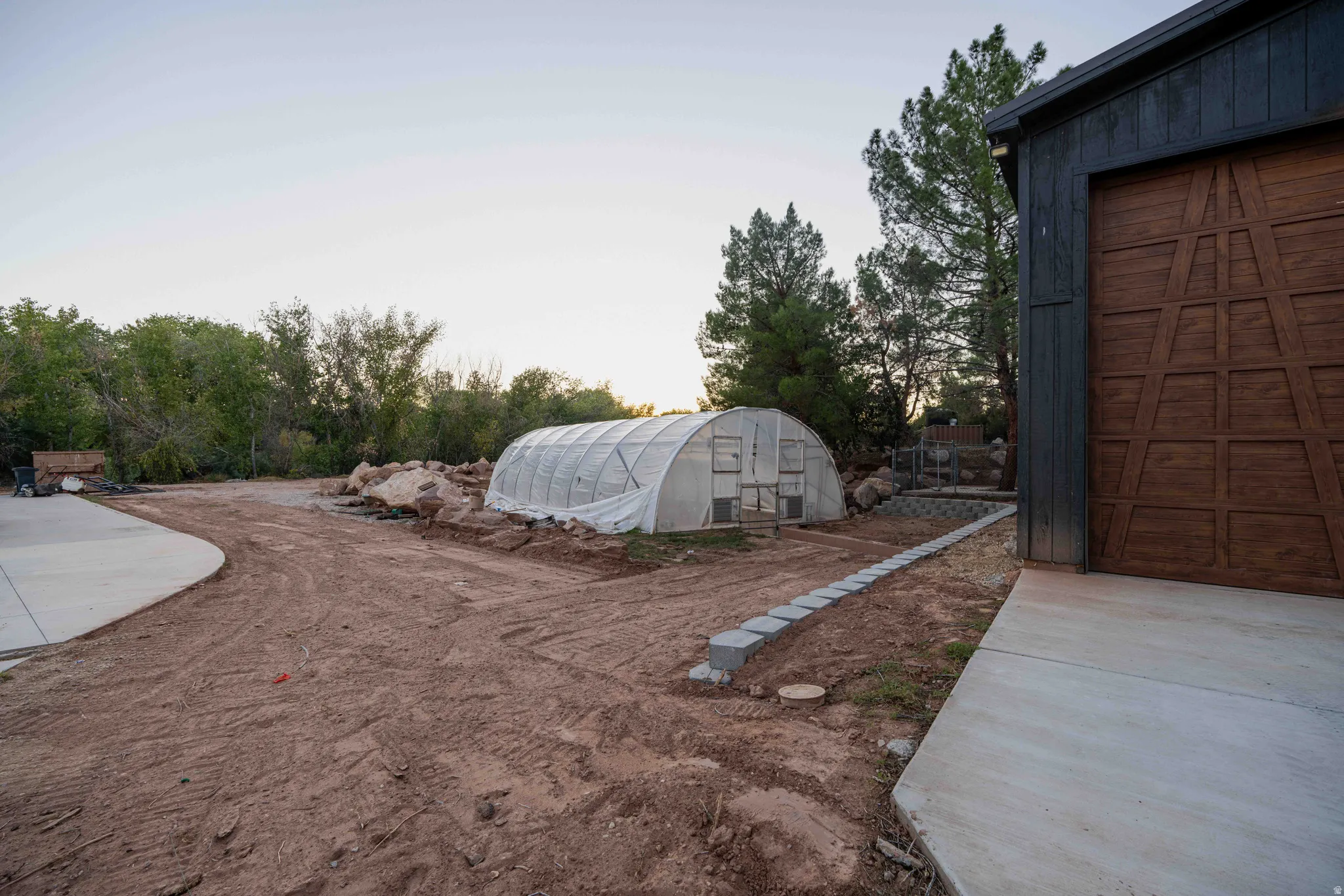 Attached garage with wood-finish door, concrete driveway, block retaining wall, and a greenhouse structure