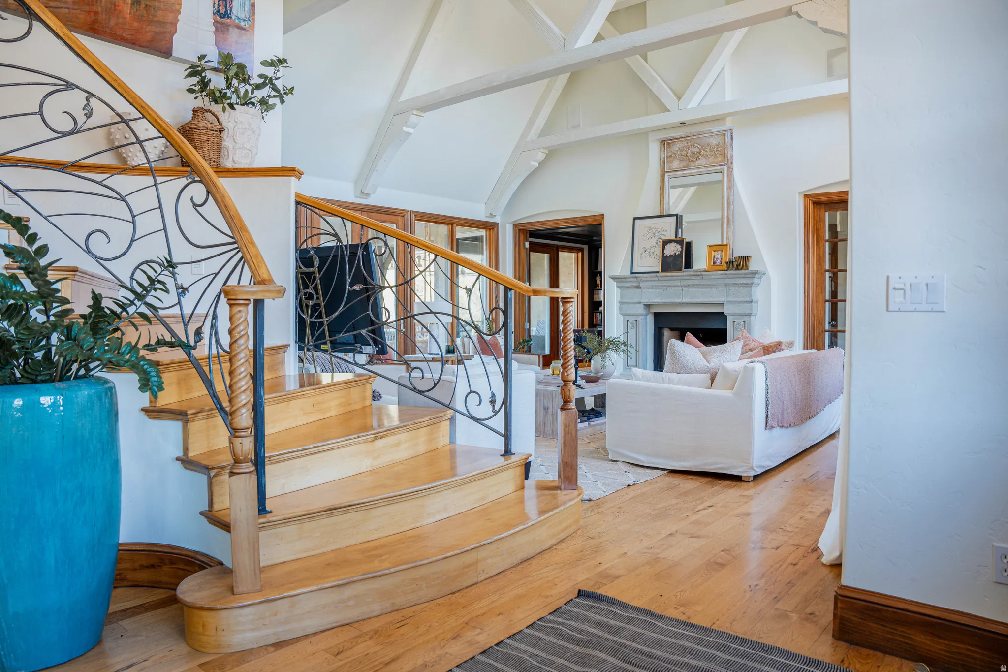 Hardwood flooring, custom wrought iron stair railing with wood handrail, vaulted ceiling with exposed beams, stone fireplace with mantel, and French doors