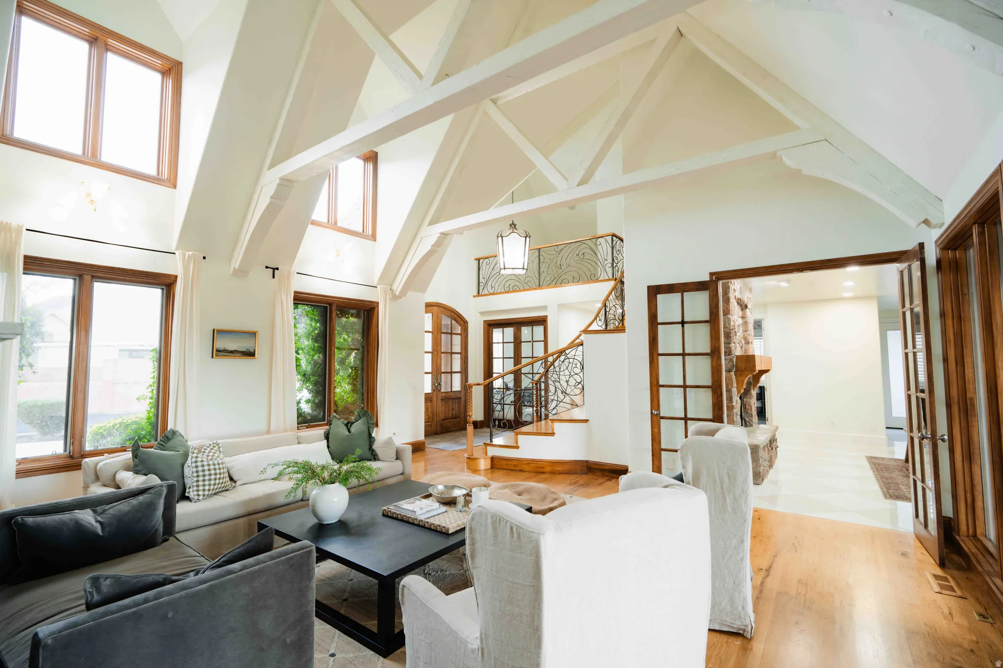 Vaulted ceilings with exposed beams, hardwood flooring, a curved staircase with wrought iron railing, French doors, and a stone fireplace