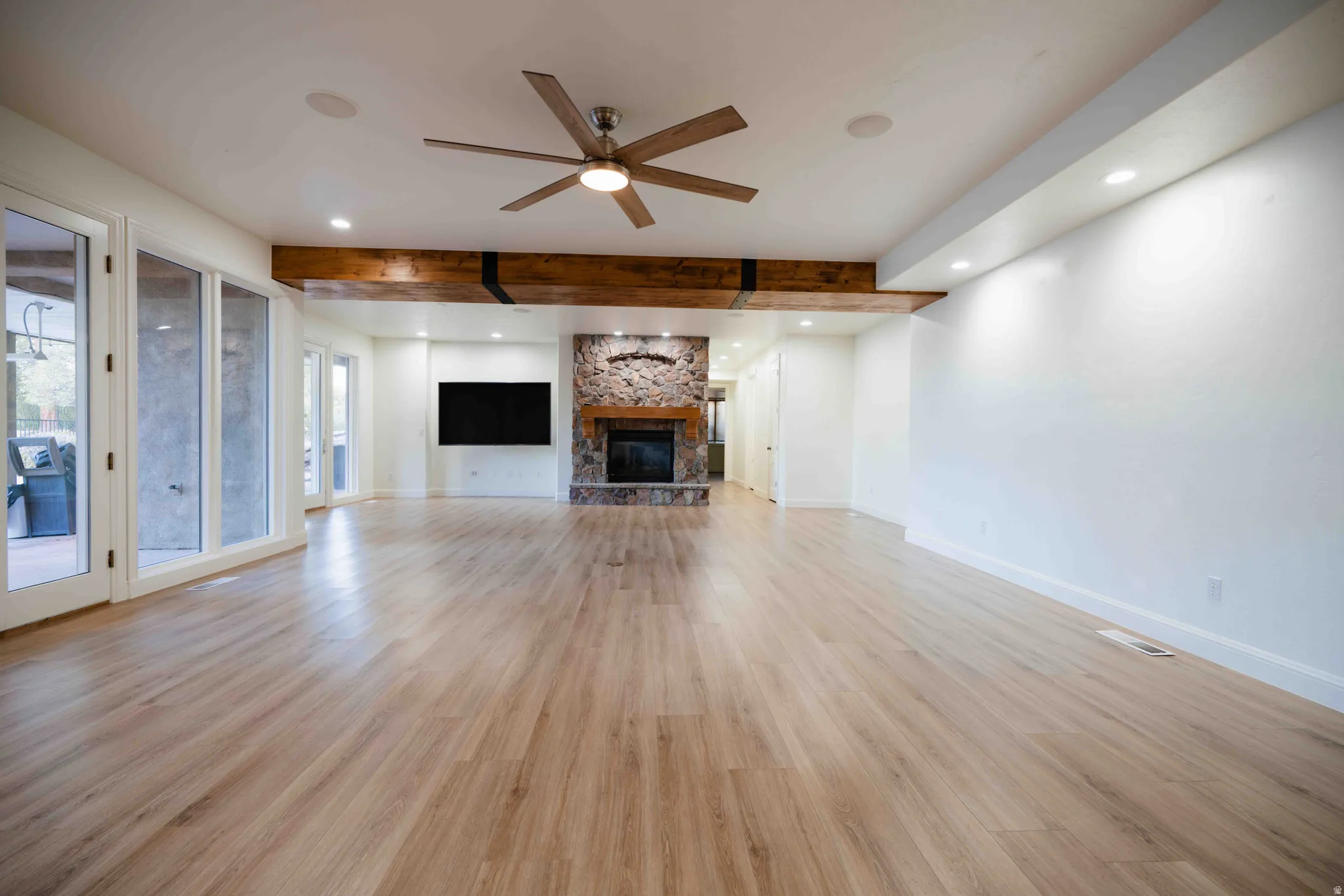 Expansive room featuring wood-finish flooring, a stone-clad fireplace, exposed wood beams, and a ceiling fan with integrated lighting