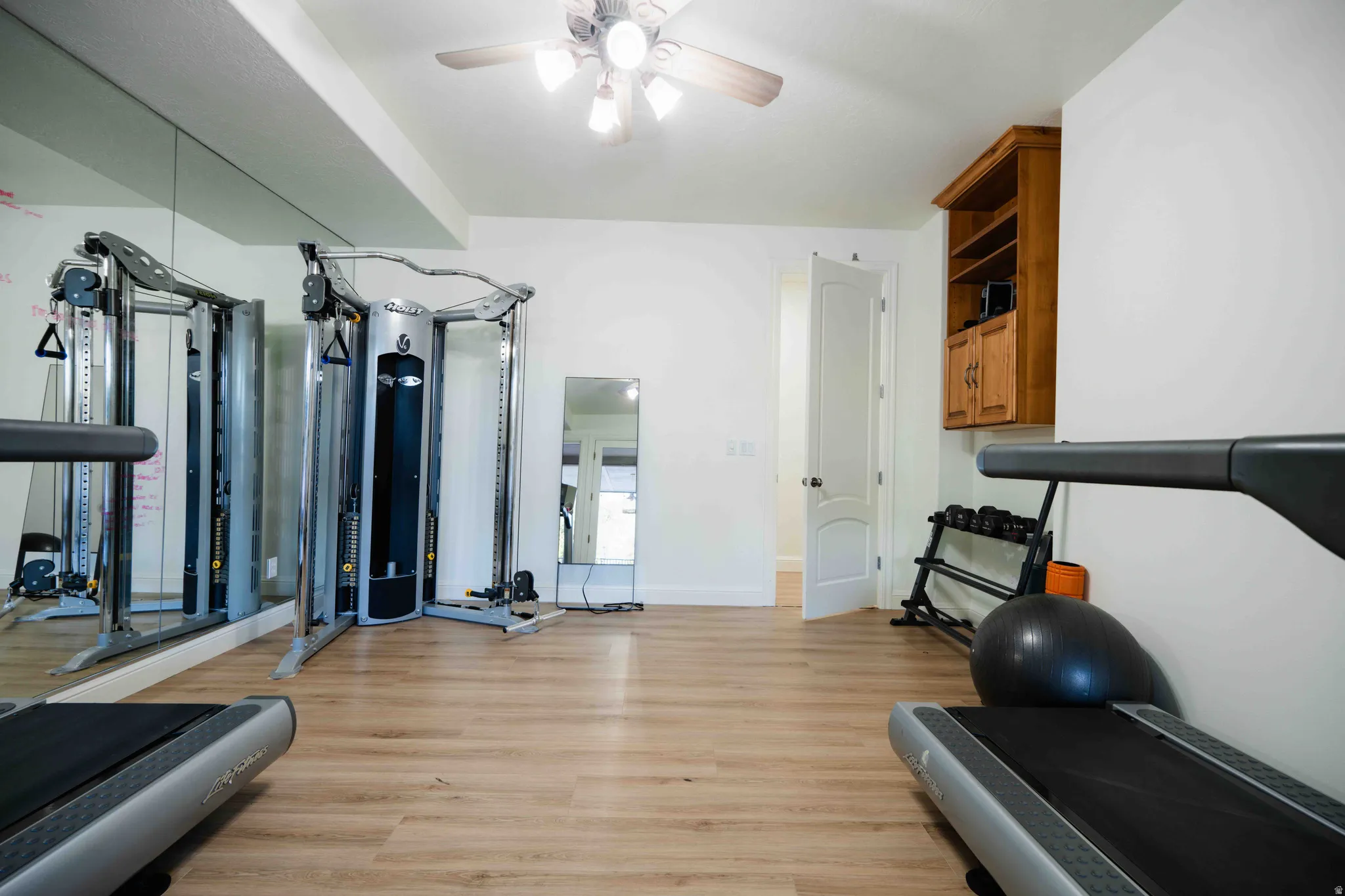 Wood-finish flooring with full wall-length mirrors, a ceiling fan with integrated lighting, a built-in wood-finish cabinet, and a five-panel interior door