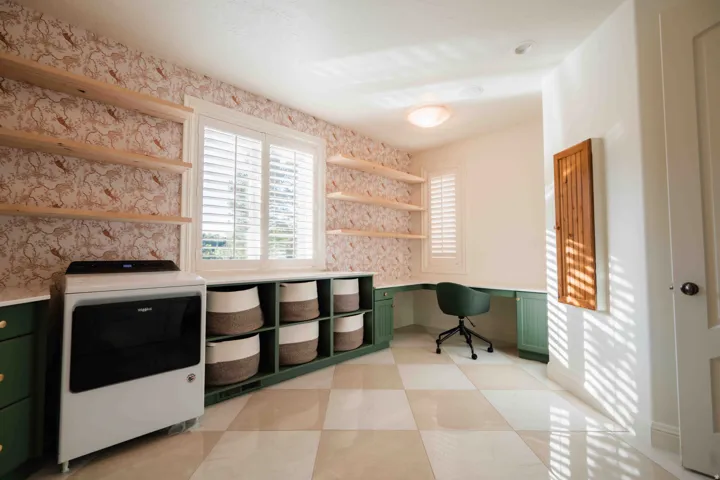 Built-in shelving, corner desk with integrated cabinetry, window with plantation shutters, patterned accent wall, and checkerboard floor tiling