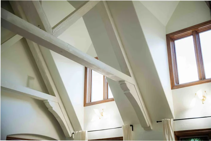 Vaulted ceiling with exposed wood beams, wood-frame windows, and wall-mounted light fixtures