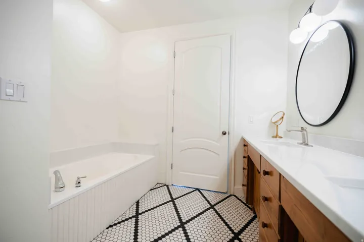 Built-in soaking tub, wood-finish vanity with dual sinks, white countertop, black-framed oval mirror, and patterned floor tile