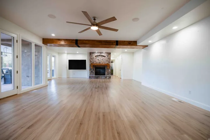 Expansive room featuring wood-finish flooring, a stone-clad fireplace, exposed wood beams, and a ceiling fan with integrated lighting