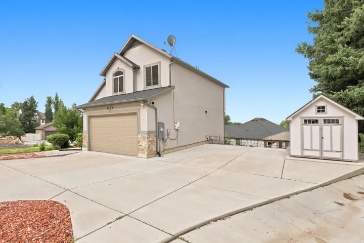 Two-story stucco exterior with stone accents, attached garage, extended concrete driveway, and a detached storage shed