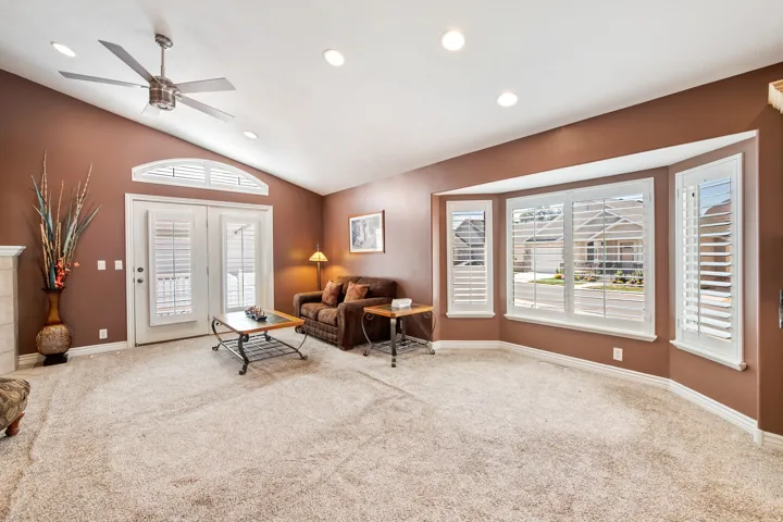 Vaulted ceiling with recessed lighting and ceiling fan, presenting a bay window and French doors with louvered shutters, and light-toned carpet flooring.