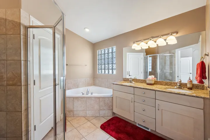 Dual vanity with integrated sinks and granite countertop, corner jetted soaking tub, glass block window, and separate glass-enclosed shower.