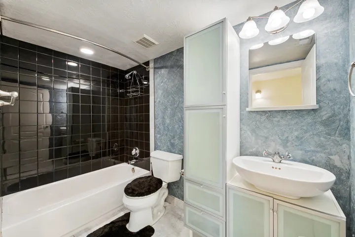 Recessed lighting, black tile shower surround, contemporary vessel sink, white vanity  and a built-in storage cabinet with frosted glass doors.