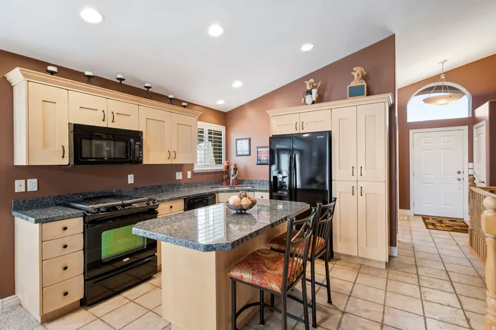 Kitchen featuring light wood-finish cabinetry, black appliances, a central island with granite countertop, and bar seating.