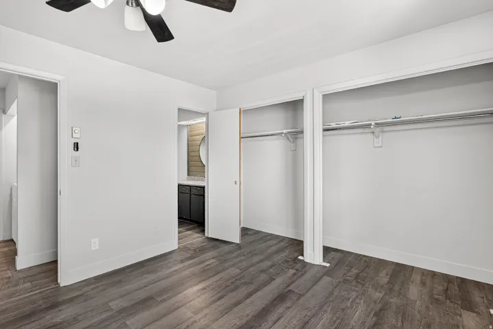 Wood-finish flooring, ceiling fan, white walls, built-in closet storage, and a partial bathroom with a vanity and round mirror
