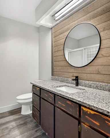 Granite countertop, wood-finish flooring, wood plank accent wall, round mirror, and a single vanity with dark cabinetry