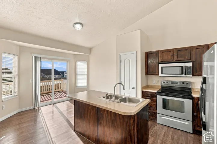 Kitchen featuring a center island with sink, wood-finish flooring, dark wood cabinetry, stainless steel appliances, and a sliding glass door to an exterior deck