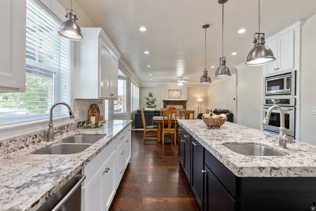 Kitchen featuring dual undermount sinks, stone countertops, white upper and lower cabinetry, a built-in oven and microwave, and dark wood-finish flooring